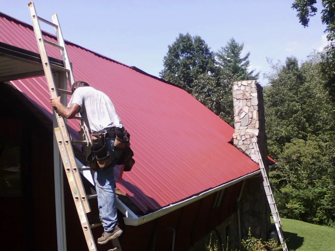 Roofer on a ladder beside a red metal roof and stone chimney — Soffit & Fascia Repair by Roofing Brothers in Ralph